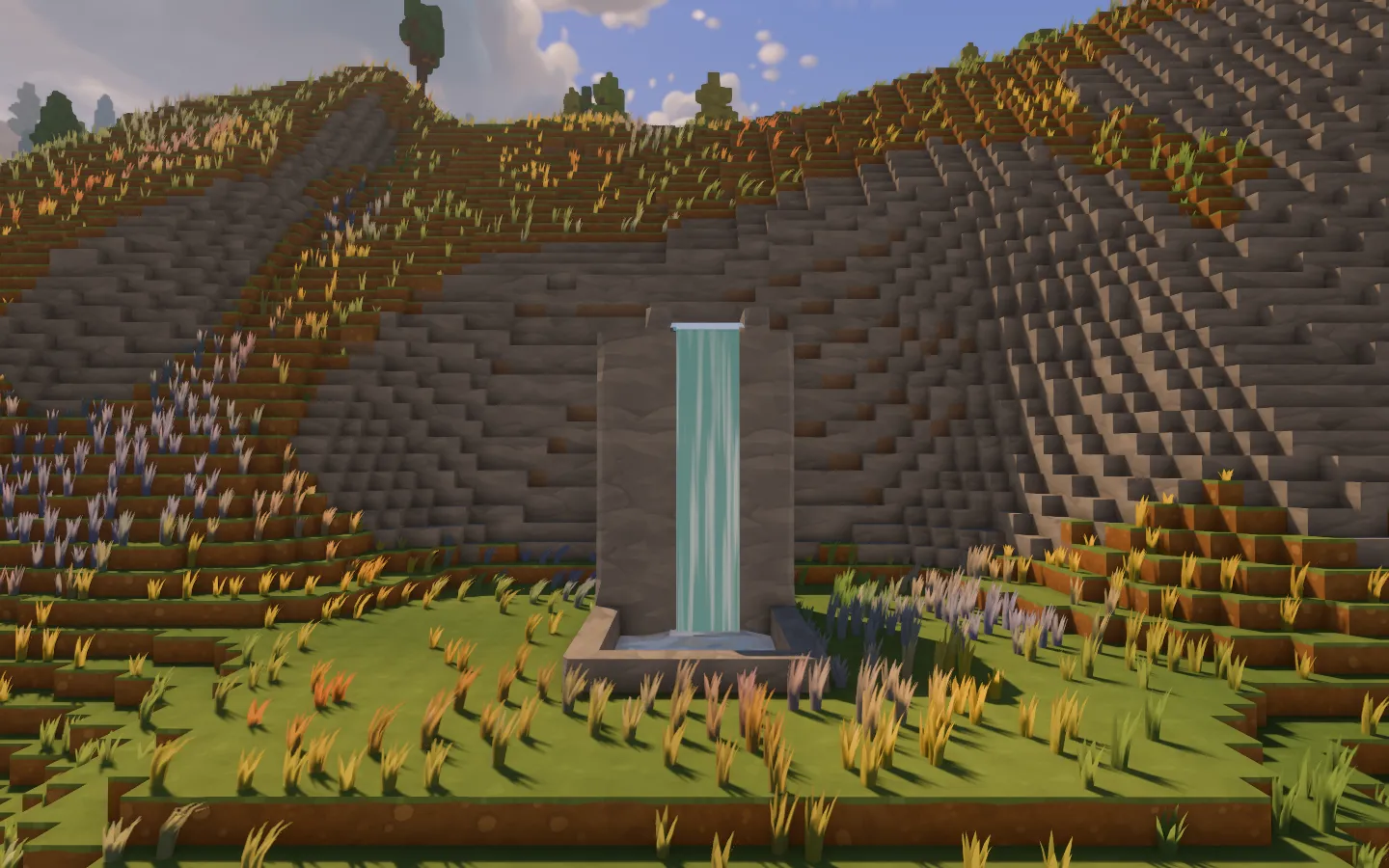 A narrow turquoise waterfall pouring between stone sidewalls into a pool at the foot of a stepped, grassy hillside.